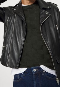 Zwarte leren bikerjack met zilverkleurige hardware, gelaagd over een donkergroen gestructureerd sweater en witte shirt, gecombineerd met blauwe jeans.