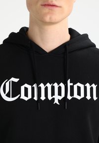 Felpa nera con cappuccio che presenta una prominente grafica bianca "Compton" in un font stilizzato, realizzata in un tessuto morbido con lacci sul cappuccio.