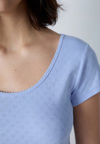 Top de color azul claro con escote redondeado, mangas cortas y tejido texturizado con un patrón de corazones. Bordes ondulados a lo largo del escote.