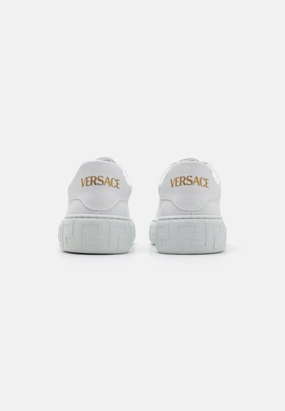 Vita läderskor med gummisula, med guld "VERSACE"-logotyp och ett mönstrat häl, som visar en stilren, minimalistisk design.