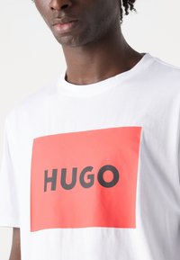 Hvit bomull t-skjorte med et stort rødt rektangulært grafikk med "HUGO" i fete svarte bokstaver. Rund hals og korte ermer.