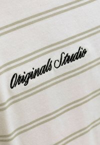 Κοντινή λήψη υφάσματος με μπεζ και άσπρες ρίγες, με μαύρο κεντημένο κείμενο «Originals Studio» σε καλλιγραφική γραμματοσειρά.