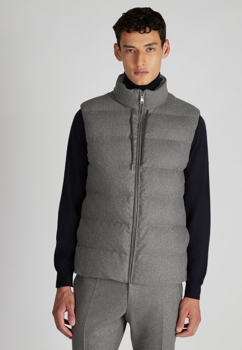 Graue gepolsterte Weste mit hohem Kragen, Frontreißverschluss und zwei Seitentaschen, über einem marineblauen Langarmoberteil getragen. Strukturierter Stoff mit leichtem Glanz.