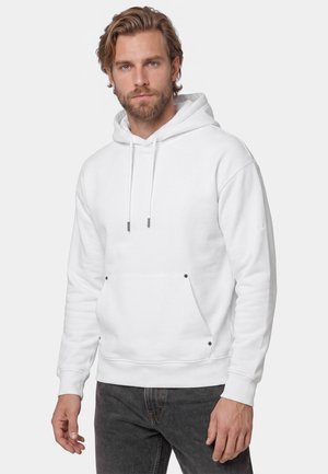 Man with light brown hair and beard wearing a plain white hoodie and dark jeans, standing against a plain light background.