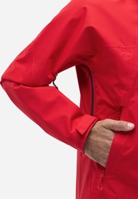 Veste imperméable rouge avec une surface texturée, dotée d'une poche latérale zippée et de poignets ajustables. Bras positionné pour accéder à la poche.