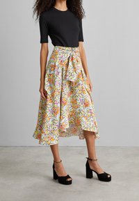 Jupe midi fleurie avec un fond bleu clair, ornée de fleurs orange et roses, une ceinture nouée à la taille et un ourlet asymétrique. Sandales à talons noires.