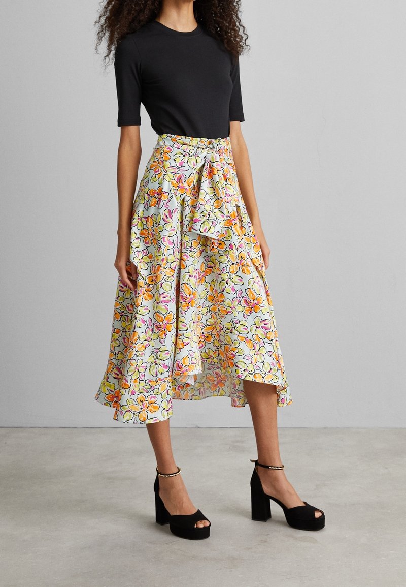 Jupe midi fleurie avec un fond bleu clair, ornée de fleurs orange et roses, une ceinture nouée à la taille et un ourlet asymétrique. Sandales à talons noires.