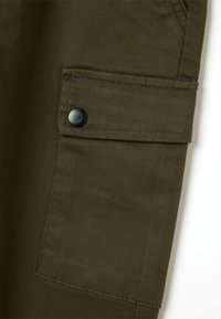 Olivegrüner Cargo-Hosenstoff mit einer großen Seitentasche, die mit einem schwarzen Druckknopfverschluss und sichtbaren Nahtdetails versehen ist.