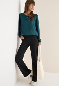 Türkise Bluse mit langen Ärmeln und Rundhalsausschnitt, schwarze Hose und weiße Sneakers. Beiger gesteppter Schultertasche. Einfaches Design, glatte Textur.