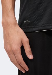 Gros plan sur une main à côté de l’ourlet d’un t-shirt de sport noir avec le texte "DRYCELL" sur le tissu.
