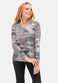 Vrouw met blond haar, gekleed in een langmouw patroon top in grijs, wit en mauve tinten, gecombineerd met zwarte broek, staand met de handen samengevouwen.