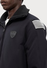Črn zip-up jopič z teksturiranim ovratnikom, gumijasto oznako in belimi črtami na rami. Gladka tekstura tkanine.