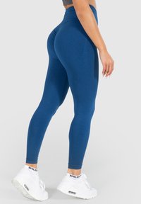 Hochtaillierte, figurbetonte Leggings in Dunkelblau, aus dehnbarem Material mit glatter Textur. Kombiniert mit weißen Sportschuhen.