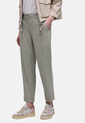 Person trägt beige Jacke, weißes Hemd, graue, zulaufende Hosen mit elastischem Bund und weiße Turnschuhe mit pinkfarbenen Akzenten, steht mit einer Hand in der Tasche.