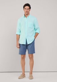  lichtblauwe linnen overhemd met knoopsluiting en een enkele borstzak, gecombineerd met donkerblauwe shorts en beige instapschoenen.