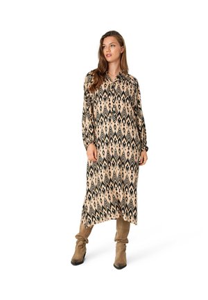 Pitkä, beige mekko, jossa on musta geometrinen kuvio, varustettuna kauluksella ja pitkillä hihoilla, yhdistettynä polven yli ulottuviin beigeihin saappaisiin.