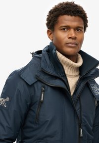 Chaqueta aislante de color azul marino con capucha con cremallera, cuello acanalado y múltiples bolsillos con cremallera. La tela tiene una sutil textura acanalada.