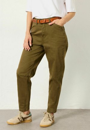 Person wearing olive green tapered pants, a white shirt, brown belt, and beige sneakers standing with one hand in a pocket.