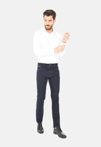 Uomo in camicia bianca a maniche lunghe che sistema il polsino, indossando jeans slim scuri e scarpe eleganti nere, in piedi contro uno sfondo chiaro e semplice.