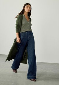 Long green cardigan over a light green button-up shirt, paired with wide-leg dark denim jeans and brown pointed-toe shoes. Plain background.