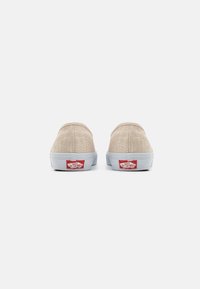 Zapatos slip-on de lona beige con una superficie texturizada, suelas blancas y un logo rojo en el talón. Punta redonda y diseño minimalista.