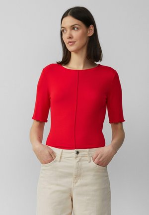 Rood geribbeld top met korte mouwen en een ronde hals, met golvende mouwen, gecombineerd met lichtbeige hoge taillebroek, handen in de zakken.