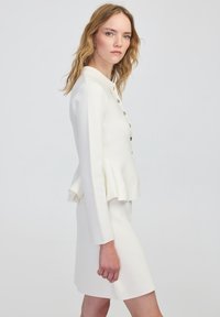 Chaqueta peplum blanca con mangas largas y cierre de botones, combinada con una falda a juego. Textura suave y corte entallado.