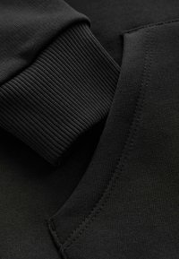 Detalle de sudadera negra que muestra un puño acanalado y una tela texturizada con una línea de costura limpia, enfatizando la simplicidad en el diseño.