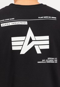 Čierne bavlnené tričko s bielym logom, textom a grafikou. Obsahuje detaily ako "ALPHA INDUSTRIES" a výrobné kódy.