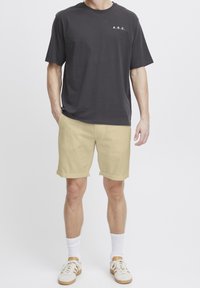 T-shirt grigia scura con logo bianco "A.R.C.", abbinata a pantaloni corti beige chiaro. Il modello indossa calzini bianchi e sneakers beige. Design semplice, vestibilità casual.