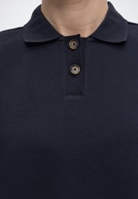 Κοντινό πλάνο ενός ατόμου που φοράει σκούρο μπλε πόλο πουκάμισο με γιακά και δύο καφέ κουμπιά κλεισμένα στο λαιμό.