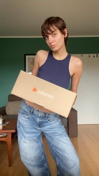 Braune Kartonschachtel mit dem "zalando"-Logo in Orange, gehalten vor einem blauen Tanktop. Der Hintergrund zeigt eine grüne Wand und einen Holzboden.