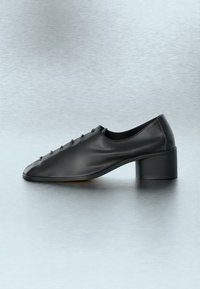 Μαύρο δερμάτινο παπούτσι με χαμηλό τακούνι, στρογγυλεμένη τετράγωνη μύτη και μικρές λεπτομέρειες που μοιάζουν με κουμπιά στην πάνω πλευρά, πάνω σε ανακλαστική γκρι επιφάνεια.
