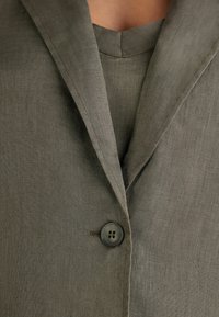 Grön linnakavaj med slagkrage, enstaka knappstängning och en strukturerad yta. Designen inkluderar en matchande innertröja.