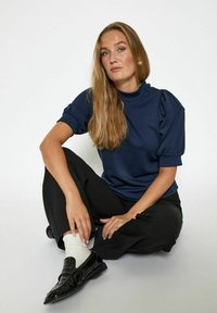 Marineblaues Kurzarmoberteil mit hohem Kragen und gerafften Puffärmeln, kombiniert mit schwarzen Hosen und glänzenden schwarzen Loafers. Weiße Socken sind sichtbar.
