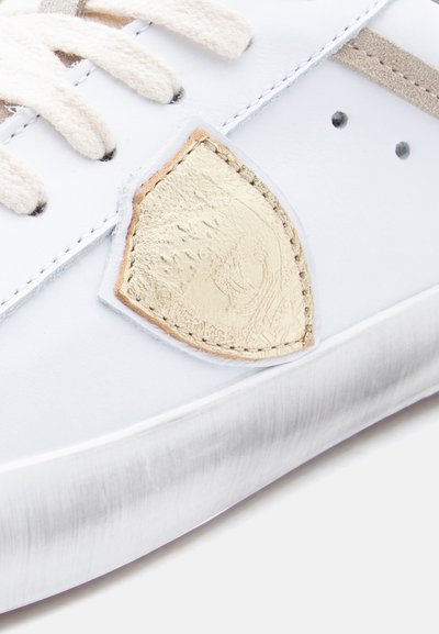 Baskets en cuir blanc avec accents beige, ornées d'un emblème doré texturé et de lacets crème. La semelle est lisse avec un léger éclat.