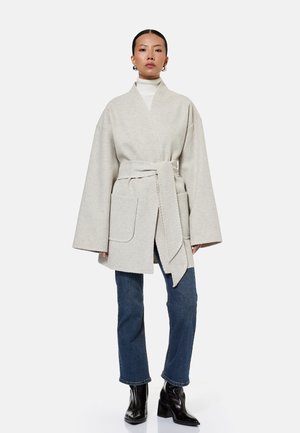 Kvinde iført en overdreven beige frakke med bælte og store lommer, hvid rullekrave, blå jeans og sorte støvler med hæl, står mod en hvid baggrund.