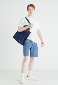 Marineblauwe totebag van stevig stof, met een bruine leren accent. Het model draagt een wit shirt, denim short en witte sneakers met sokken.