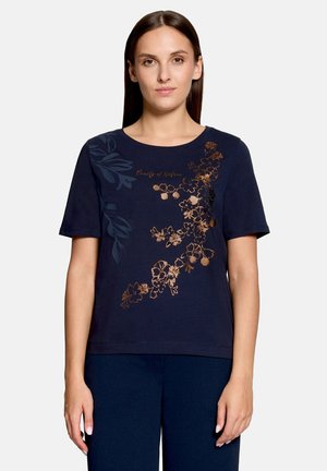 Kvinde iført en navyblå T-shirt med korte ærmer, dekoreret med et kobber- og mørkeblå blomsterprint foran og teksten "Beauty of Nature."