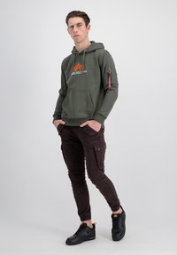 Felpa verde con tasca frontale, caratterizzata da un logo a stella arancione e una tasca con zip rossa. Pantaloni cargo marroni con polsini a coste e sneakers nere.