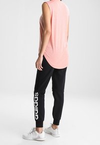 Svetle růžový top bez rukávů s zakřiveným lemem, doplněný černými joggingovými kalhotami s bílým nápisem "adidas" na boku a šedými sportovními botami.