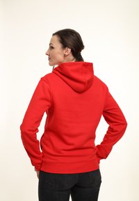 Felpa rossa con cappuccio grande, polsini e orlo a costine. Texture liscia, design aderente e silhouette rilassata. Indossata con jeans scuri.