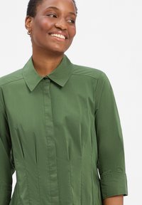 Zielona koszula z kołnierzykiem i rękawami o długości trzy czwarte, z pionowymi szwami modelującymi oraz gładką, lekką struktura materiału.