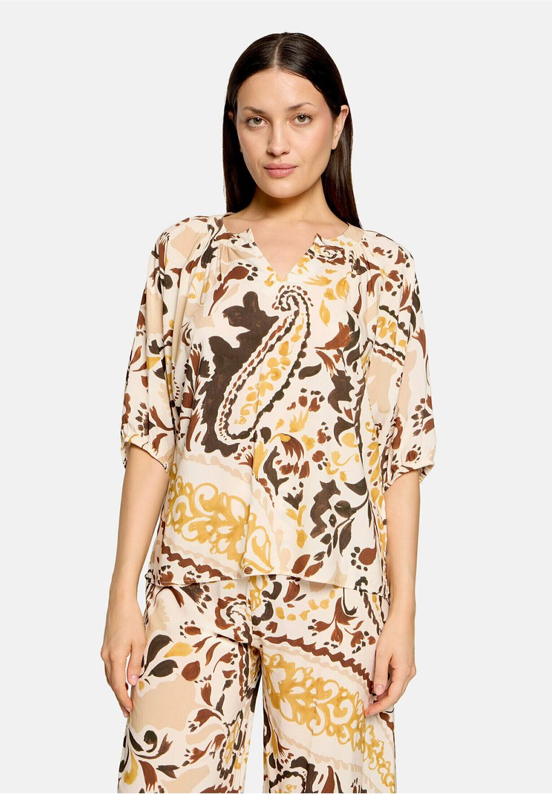 Femme portant un chemisier ample beige, marron et jaune à motif cachemire et un pantalon large assorti, debout devant un fond uni.