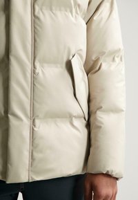 Beige pufferjack met een zachte, gladde buitenkant, voorzien van een zijzak en gestikte secties voor isolatie. Laag profiel manchet.