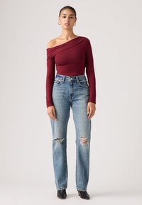 Γυναίκα όρθια με μπορντό τοπ με ένα μανίκι, σκισμένα ανοιχτόχρωμα μπλε τζιν, μαύρα μποτάκια αστραγάλου και χρυσούς κρίκους σκουλαρίκια μπροστά σε απλό φόντο.