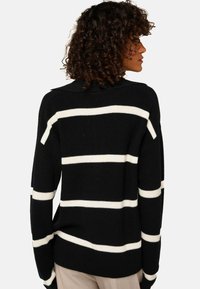 Femme aux cheveux bouclés portant un pull noir à larges rayures blanches horizontales, vue de dos, sur un fond uni.