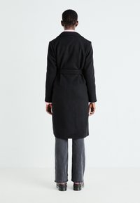 Black long coat with a tie waist, featuring a notched collar and smooth texture, worn over gray pants and chunky black shoes.