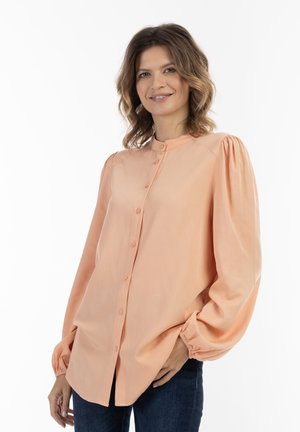 Kvinde med skulderlangt bølget hår iført en ferskenfarvet skjorte med knapper og mørke jeans, smilende med den ene hånd i lommen mod en hvid baggrund.