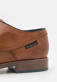 Scarpa in pelle marrone con finitura liscia, forma arrotondata, cuciture nere e una piccola etichetta con logo sul lato. Tacco in legno basso.